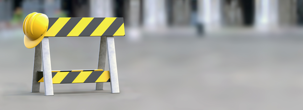 A yellow and black construction sign on a concrete floor. A yellow hard hat hangs from one corner of the sign.
