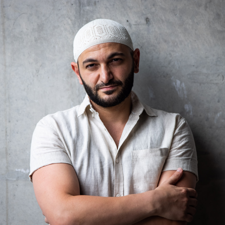Dr. Michael Mohammed Ahmed. A man wearing a light shirt and a white cap stands with folded arms gazing at camera. Image by Anna Kucera