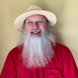 A smiling man with a long grey beard, wearing a straw hat and red shirt.