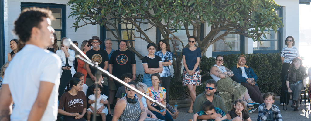 Young man talking in microphone in front of a crowd of people sitting in the sunshine outside the Whibayganba cottages.