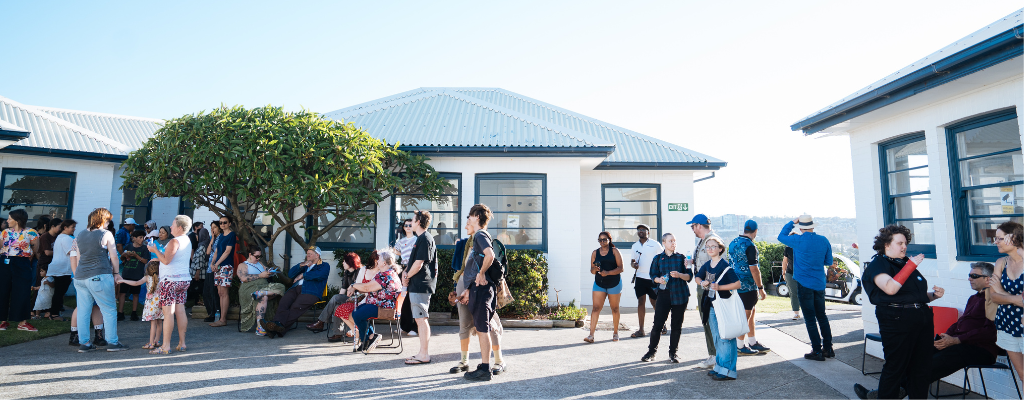 Young man talking in microphone in front of a crowd of people sitting in the sunshine outside the Whibayganba cottages.