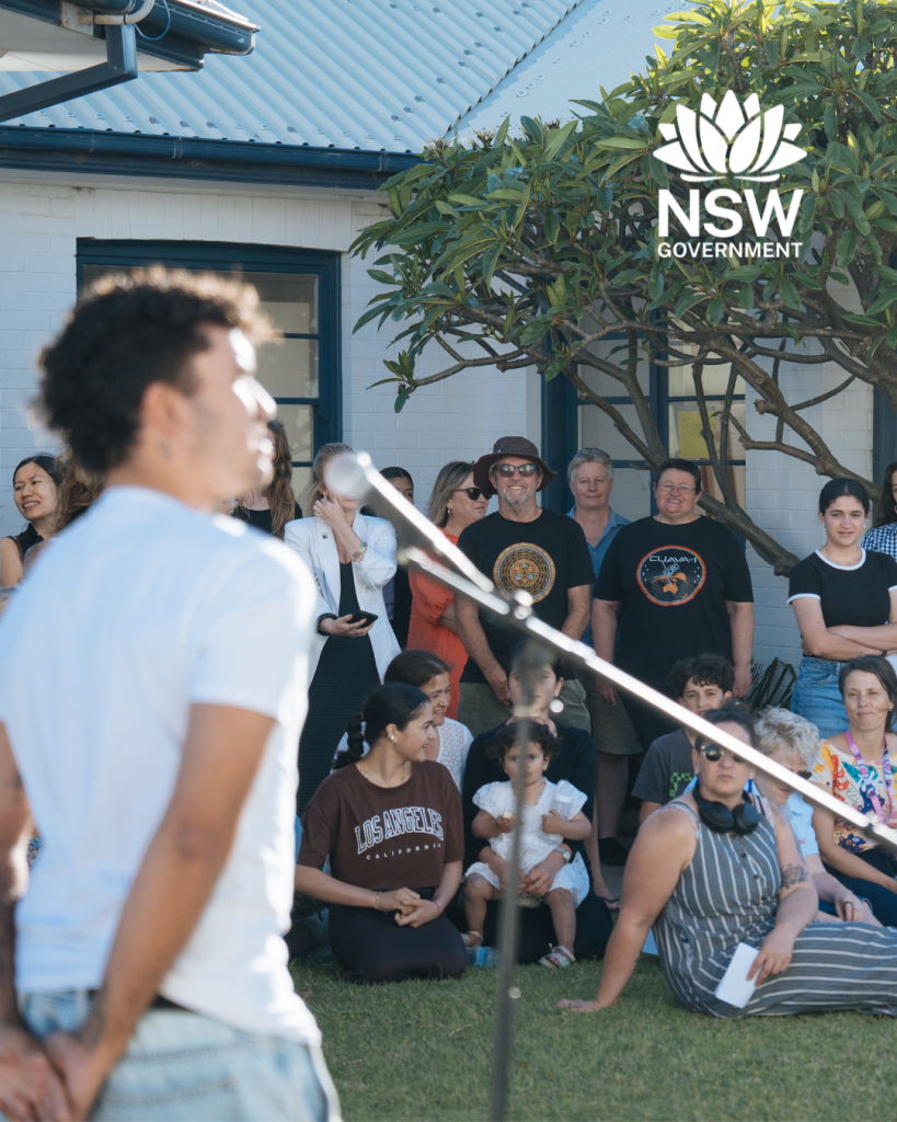 Young man talking in microphone in front of a crowd of people sitting in the sunshine outside the Whibayganba cottages.