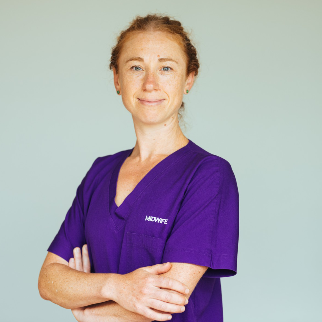 A person in a purple uniform with the word “MIDWIFE” embroidered on the chest, standing with arms crossed and smiling.