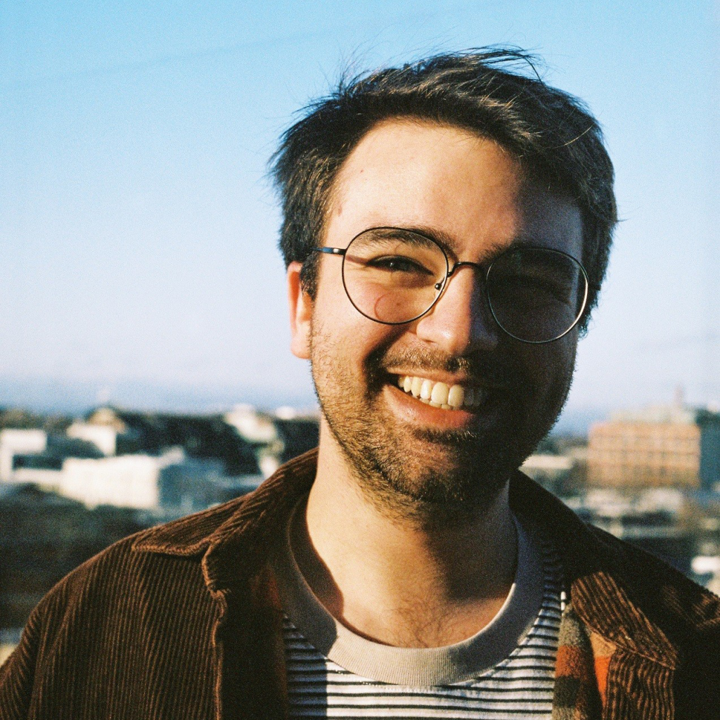 Rory Green - A smiling man with glasses wearing a striped shirt and brown jacket