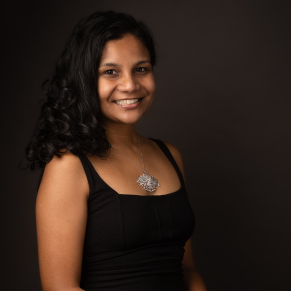 Anjalee Senarath smiling in a black top, with curled dark hair, against a dark background.