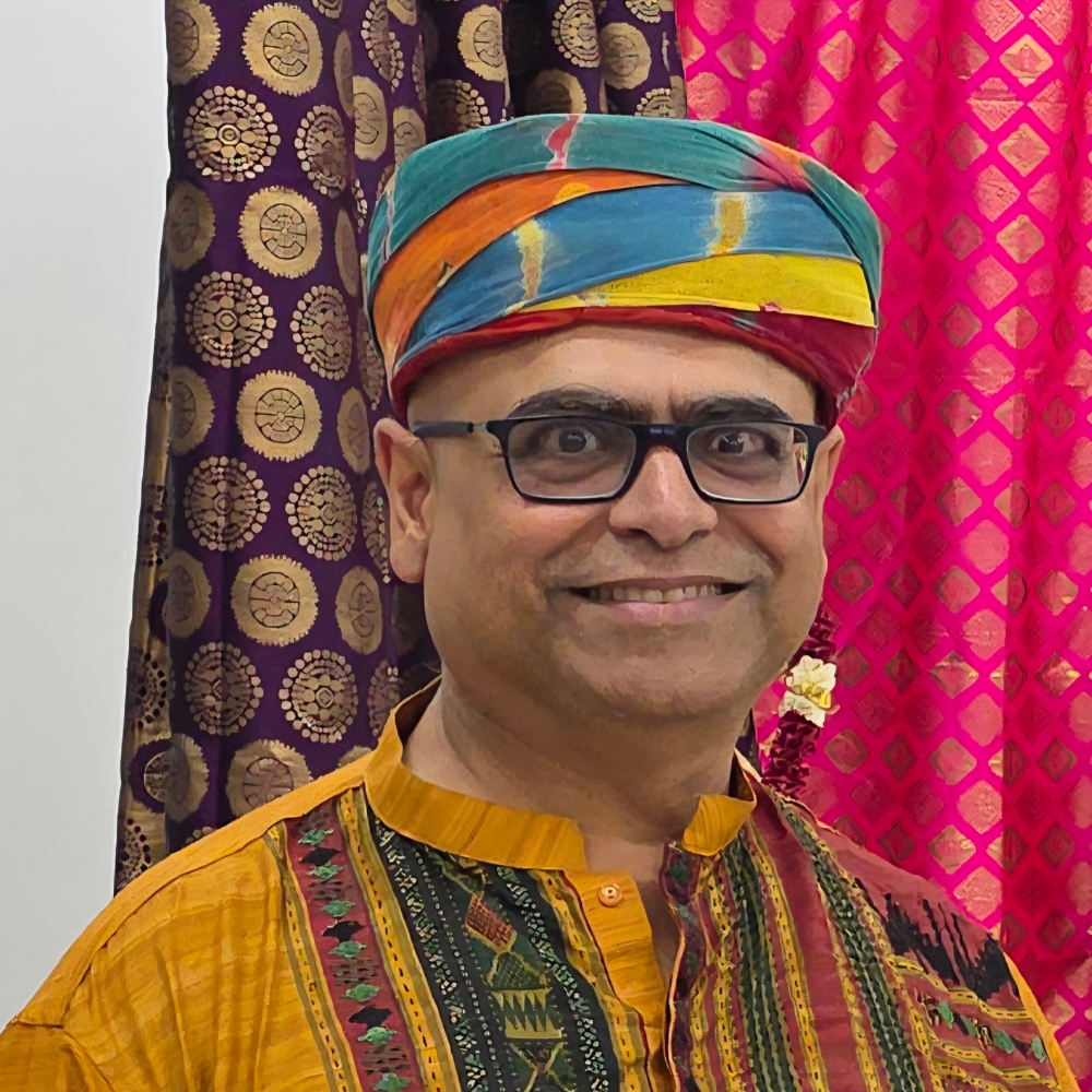 Suman Lahiry wearing glasses and a colourful traditional outfit, standing in front of patterned fabric.