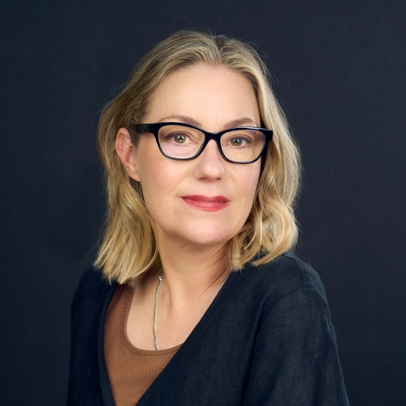 Person with shoulder-length blonde hair wearing black-framed glasses, looking slightly to the side against a dark blue background.