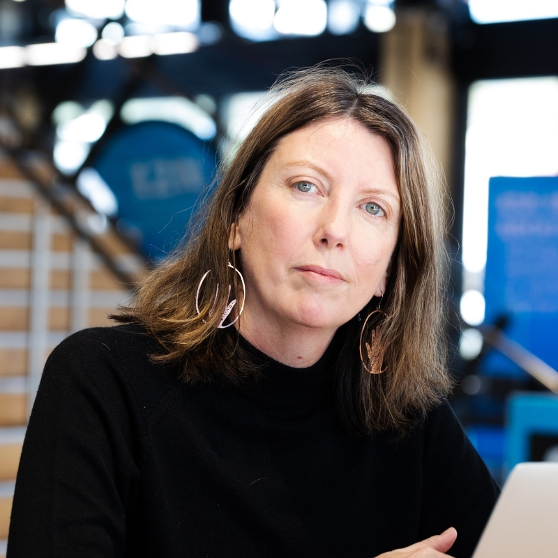 Person with shoulder-length brown hair wearing a black top and large hoop earrings, seated at a table with a laptop, looking toward the camera.
