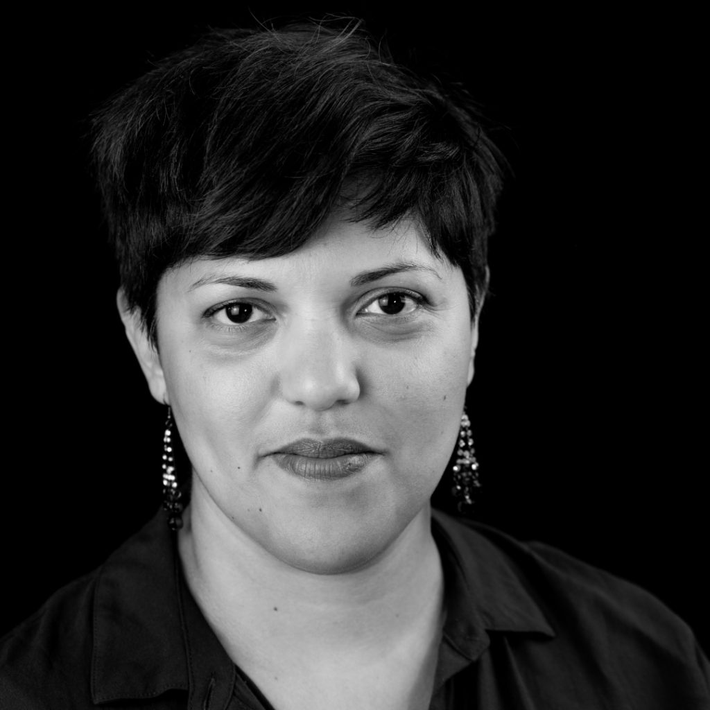Black and white headshot of a person with short dark hair wearing dangling earrings and a dark collared shirt, looking directly at the camera.